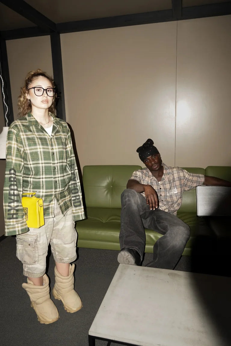 Zwei Models in grünen und beigefarbenen karierten HXYFORD-Hemden vor einer grünen Couch.