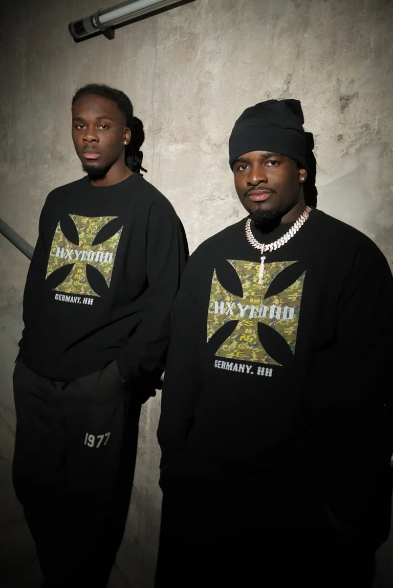Zwei Models in schwarzen HXYFORD-Sweatshirts mit grünem Grafikprint vor einer Betonwand.